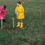 Dois voluntários plantam mudas de árvores em uma área gramada, usando capas de chuva coloridas e botas.