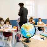 Estudantes em uma sala de aula durante uma atividade de geografia, com um globo sobre a mesa do professor.
