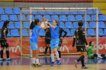 Jogadoras de futsal feminino comemoram juntas durante uma partida em quadra coberta.