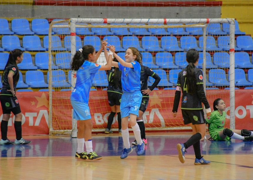 Jogadoras de futsal feminino comemoram juntas durante uma partida em quadra coberta.