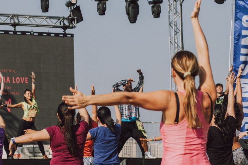Grupo de pessoas participa de uma aula de zumba ao ar livre, acompanhando os movimentos de uma instrutora.