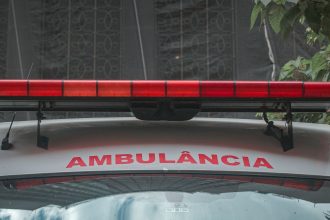 Close da dianteira de uma ambulância urbana, com a luz de emergência vermelha e a inscrição “AMBULÂNCIA”.